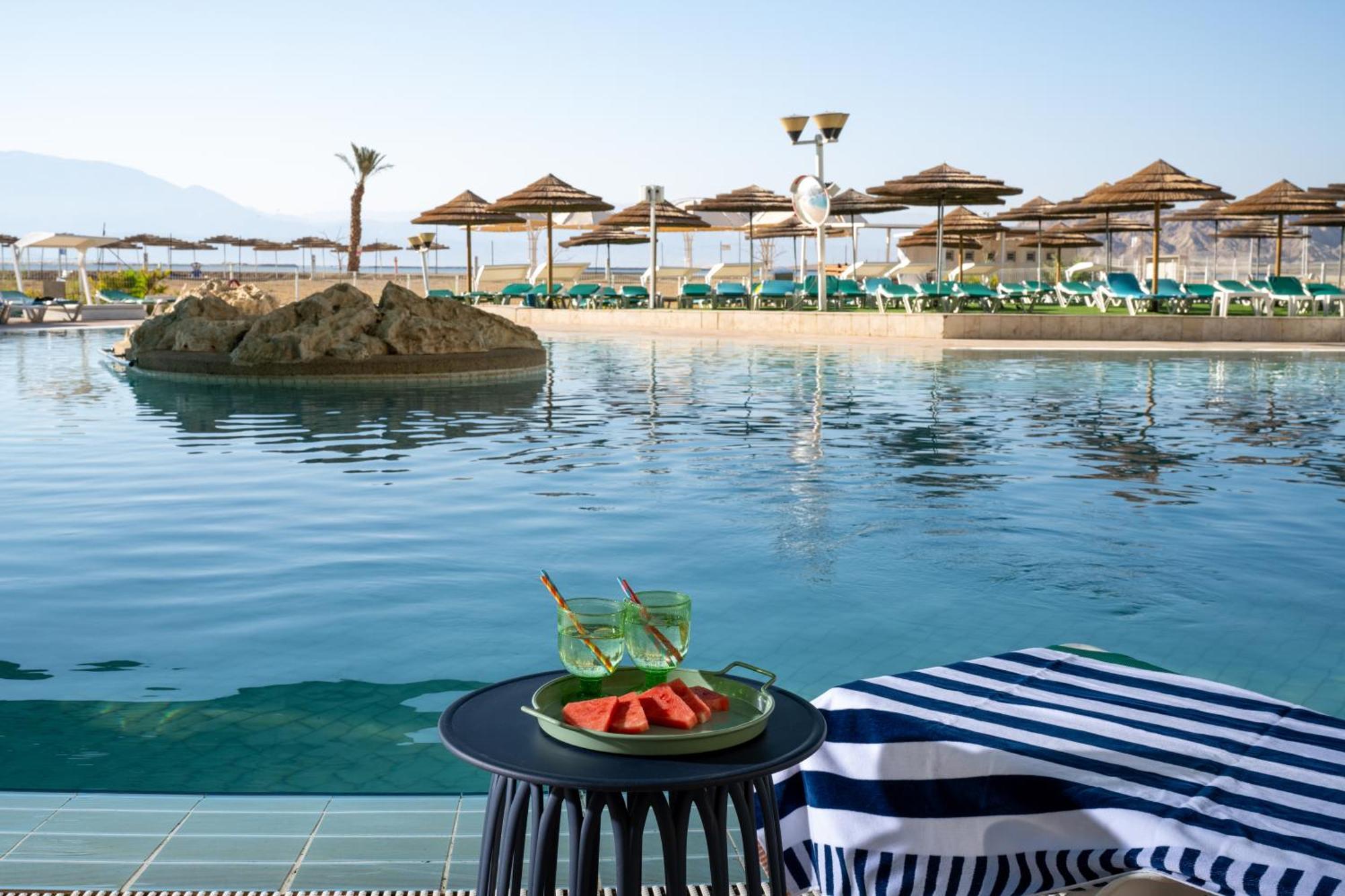Leonardo Club Hotel Dead Sea Neve Zohar Eksteriør bilde The photo depicts a tranquil swimming pool area at a resort or hotel. In the foreground, there is a small round table with a plate containing slices of watermelon and two green drinks. The background features a calm pool with a small rock island in t