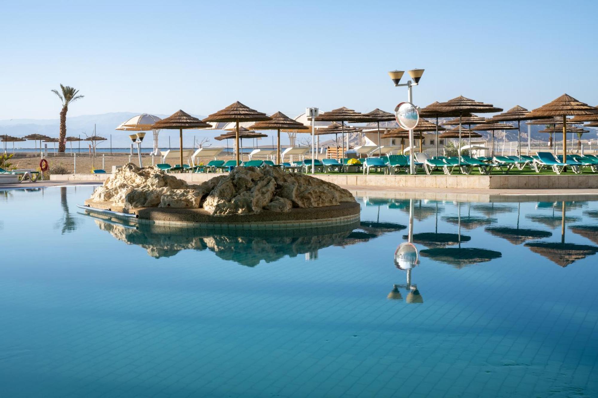 Leonardo Club Hotel Dead Sea Neve Zohar Eksteriør bilde The photo shows a tranquil swimming pool area. It features a large, reflective pool with a small rock formation in the center. Surrounding the pool are several sun loungers with green cushions and a series of thatched umbrellas providing shade. In th